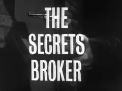 title card: white all caps text reading ‘THE SECRETS BROKER’ superimposed on a dark close-up of Paignton taking the revolver from the box