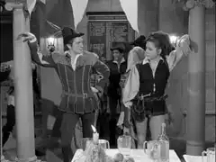 Mrs. Peel leans against a pillar, resplendent in her Robin Hood outfit and critical of Duboys for wearing the same costume; three students in mediæval costume are huddled behind her on the left