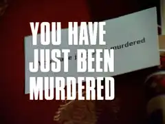 title card: white all caps text with thin black dropshadow to the right reading ‘YOU HAVE JUST BEEN MURDERED’ superimposed on the business card bearing the same text lying on the leather-topped desk